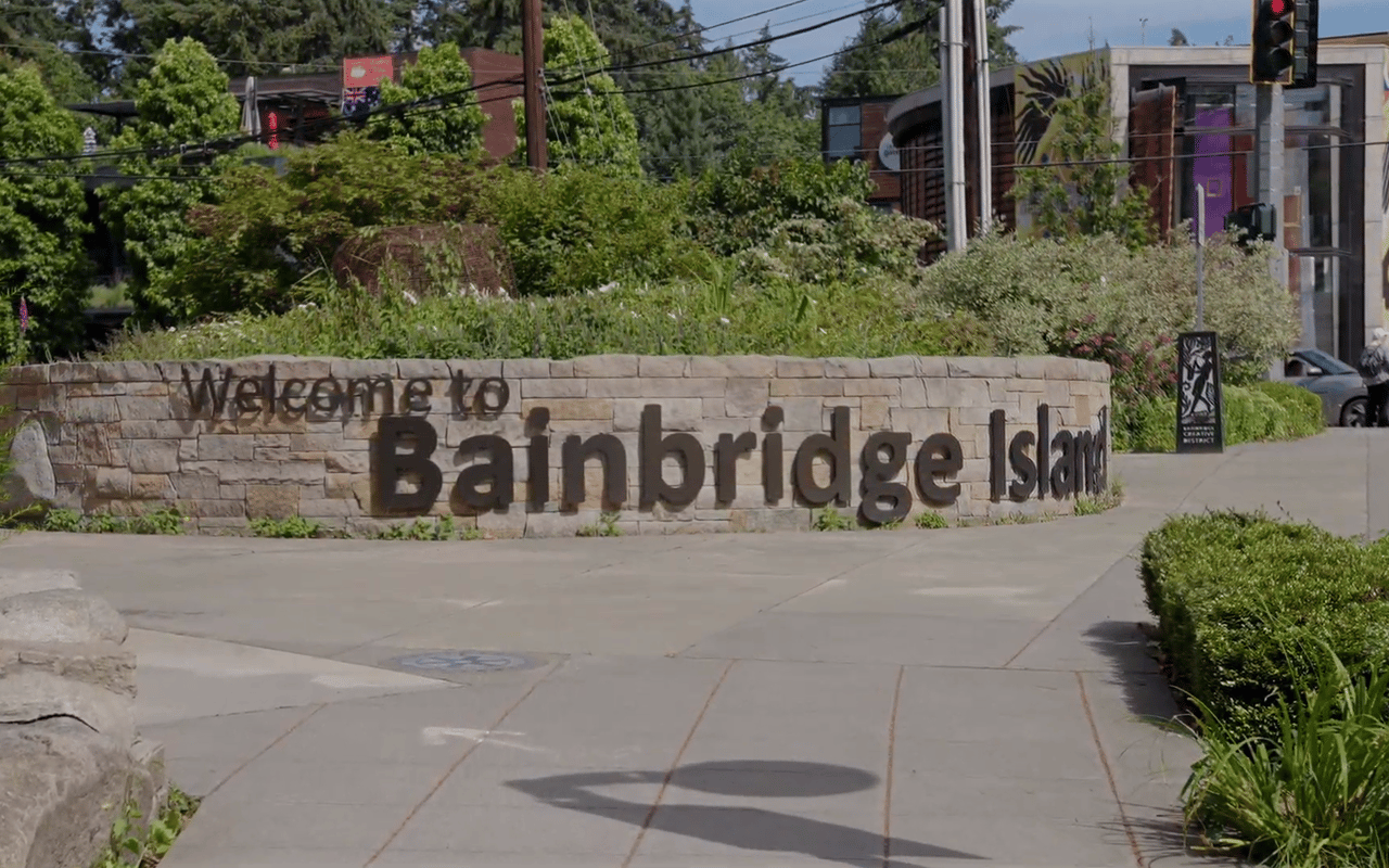 Bainbridge Island welcome sign beside a landscaped sidewalk and modern storefronts.