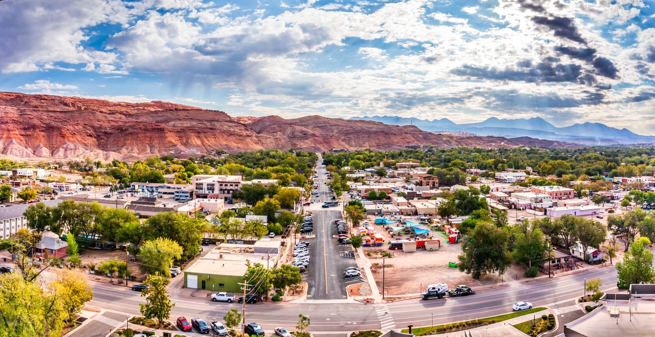 Panoramic town view with a main street, leafy trees, and red sandstone cliffs beyond.