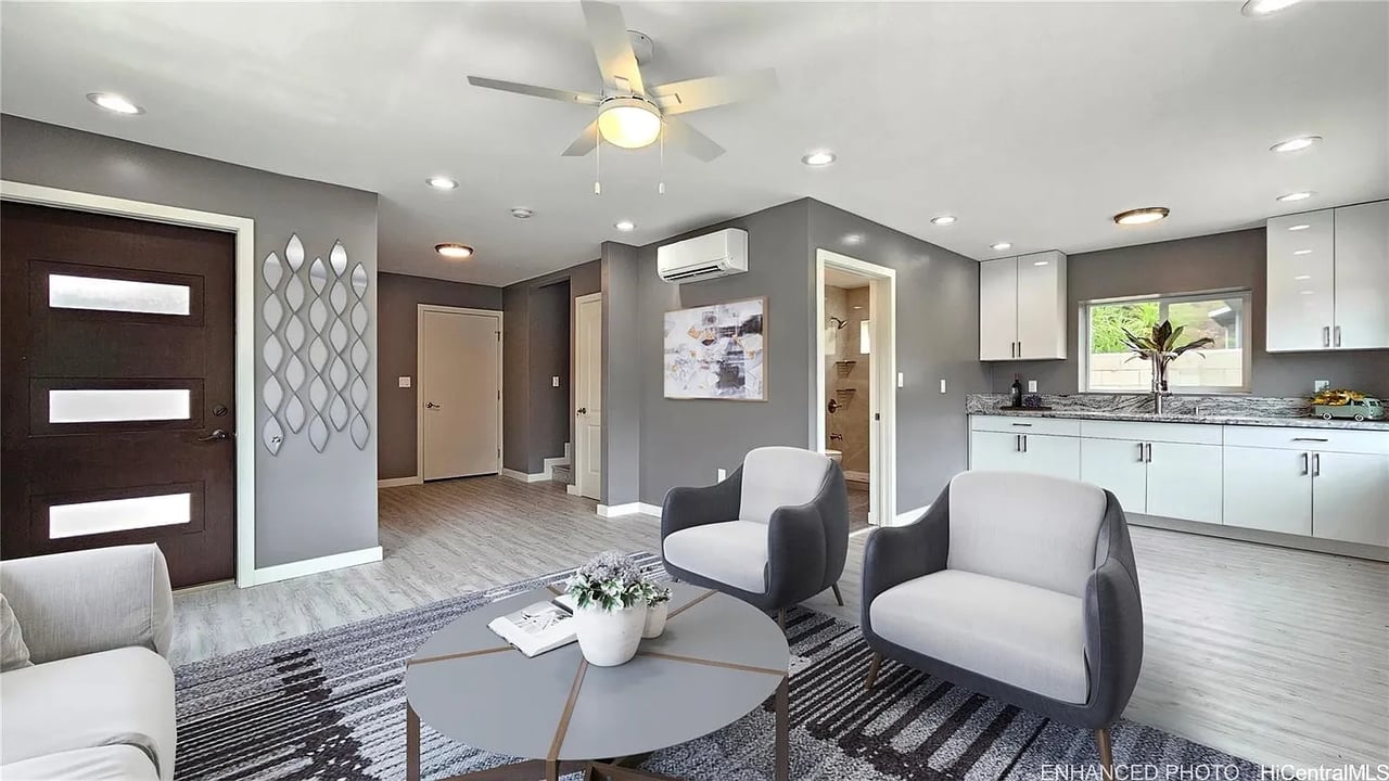 Open-plan living room and kitchen with gray armchairs, a geometric rug, and white cabinetry.