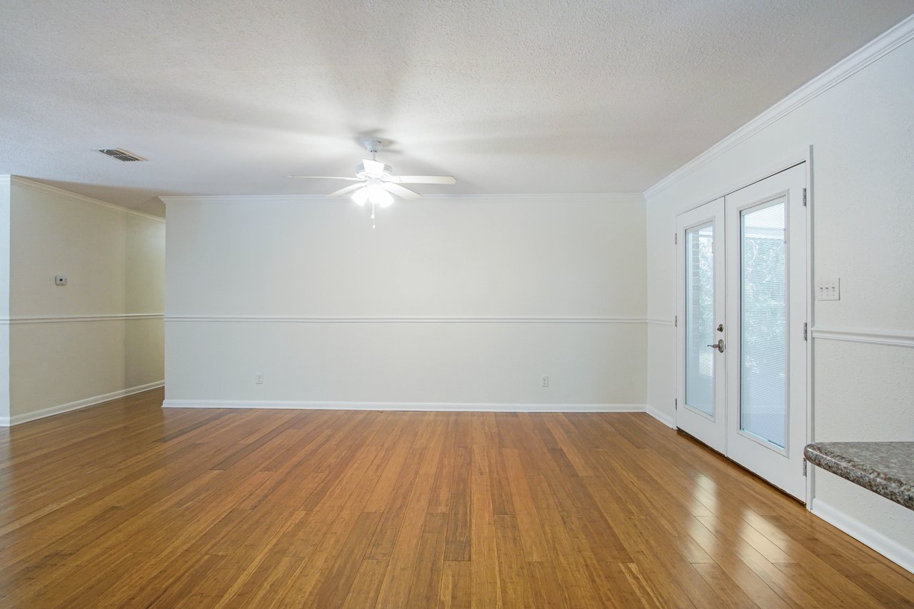 Empty room with polished wood floors, beige walls, and crown molding. A ceiling fan with light hangs from the ceiling. Double glass doors on the right.