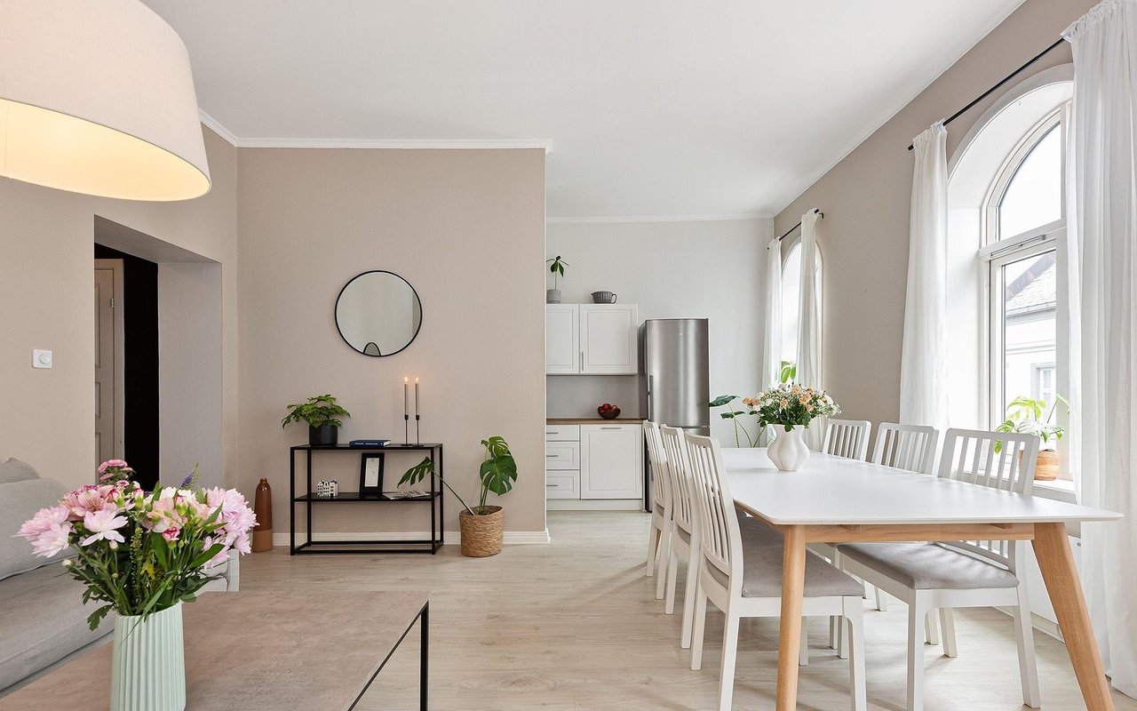 Open-plan dining area with a white table beside tall arched windows and an adjoining kitchen.