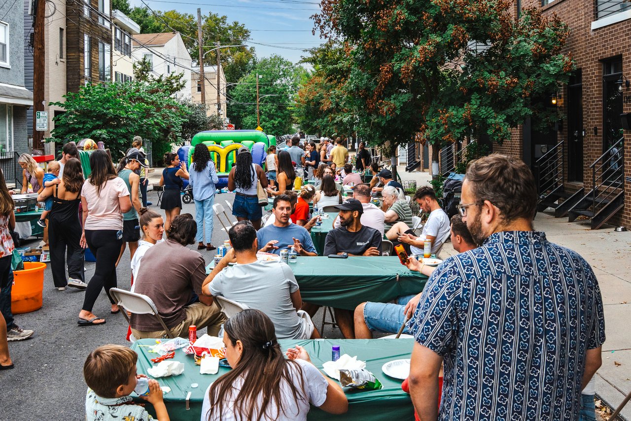 Neighbors gather at picnic tables on a residential street, with a colorful inflatable bounce house nearby.