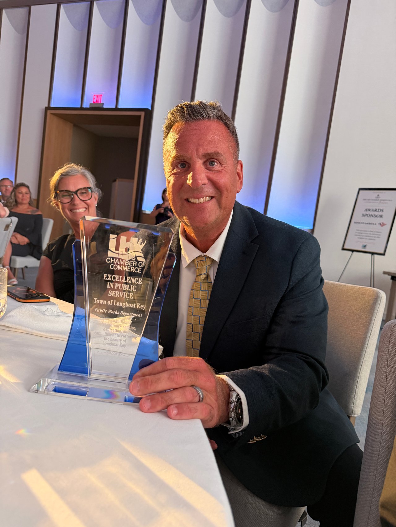 Man in a dark suit holding a Longboat Key Chamber of Commerce award beside a smiling woman.
