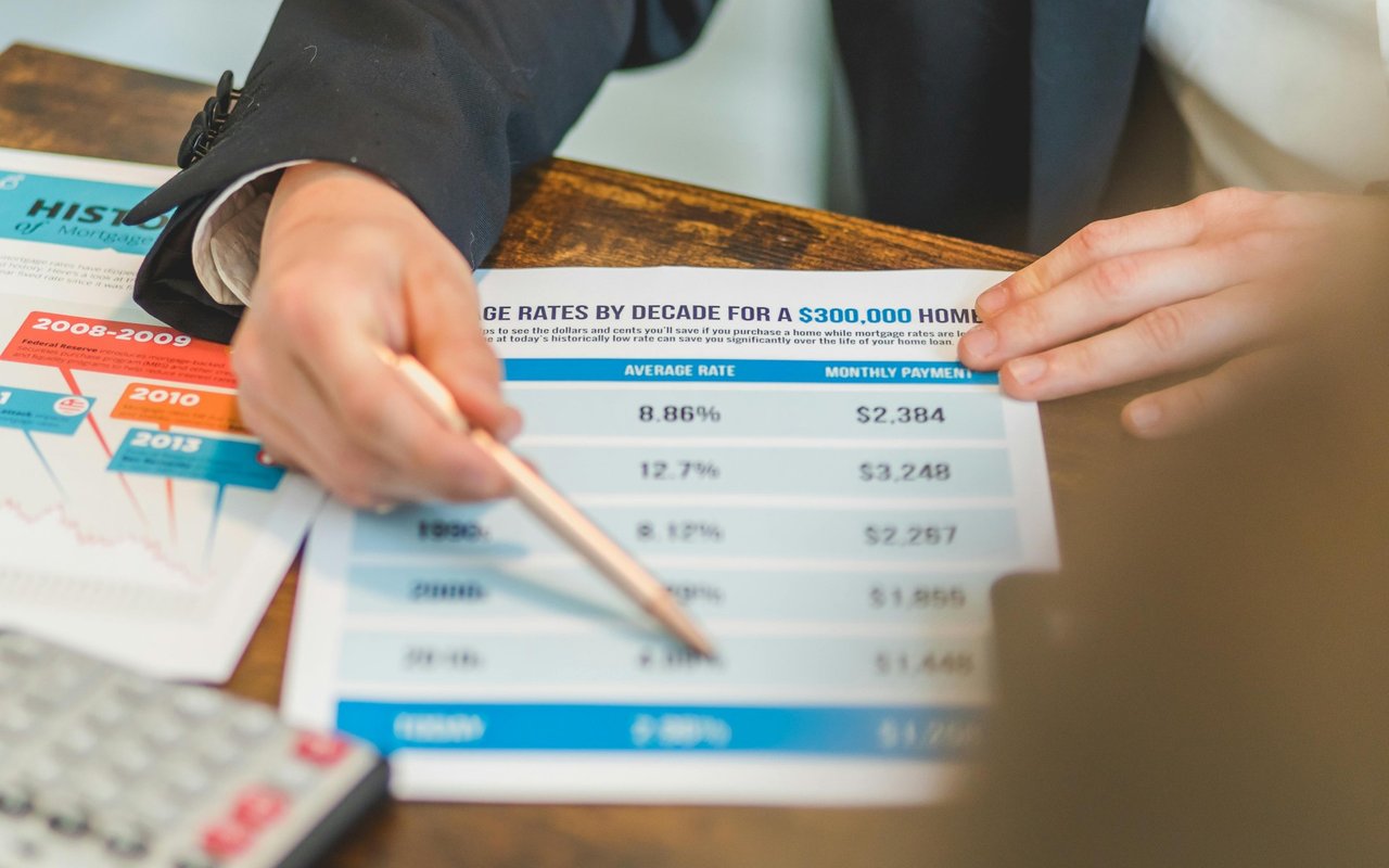 Hands point to a mortgage-rate comparison sheet beside historical rate charts and a calculator.