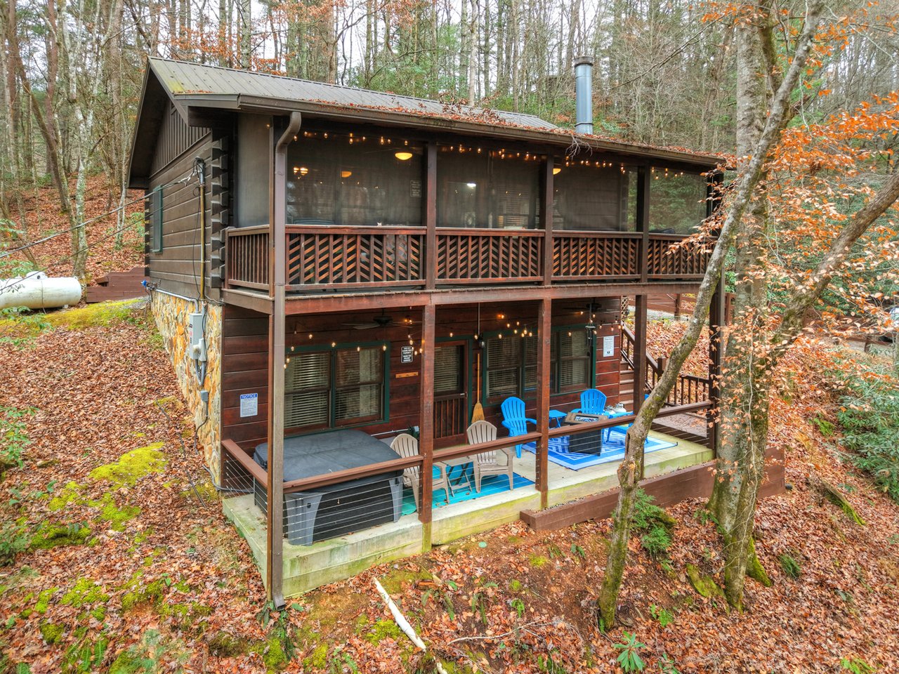 Two-story log cabin in autumn woods with a screened porch and furnished lower deck.
