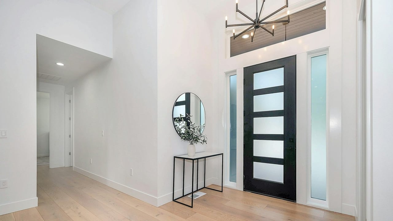Bright, empty home entryway with wide-plank wood floors, a black glass-panel front door and a round mirror over a console table