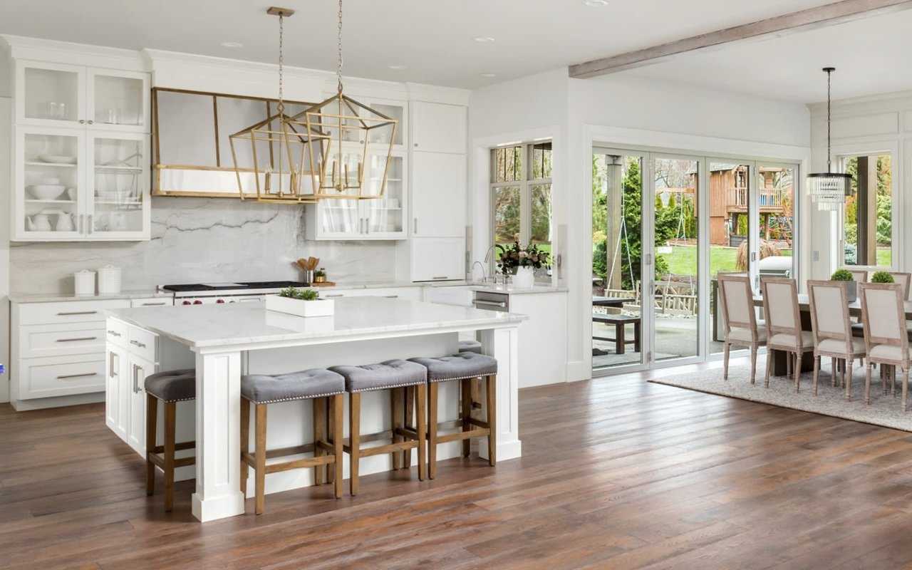 Bright white kitchen with a marble island, gray bar stools, gold pendants, and sliding doors to the garden.