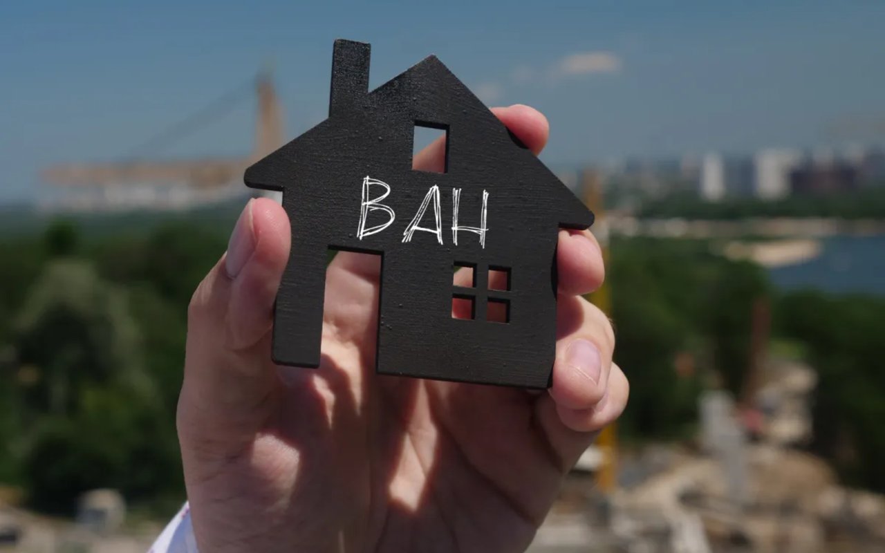 A hand holds a black house-shaped cutout marked “BAH” against a blurred city skyline.