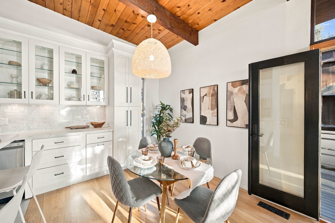Dining area beside a white kitchen, with a glass table beneath a vaulted wood ceiling.