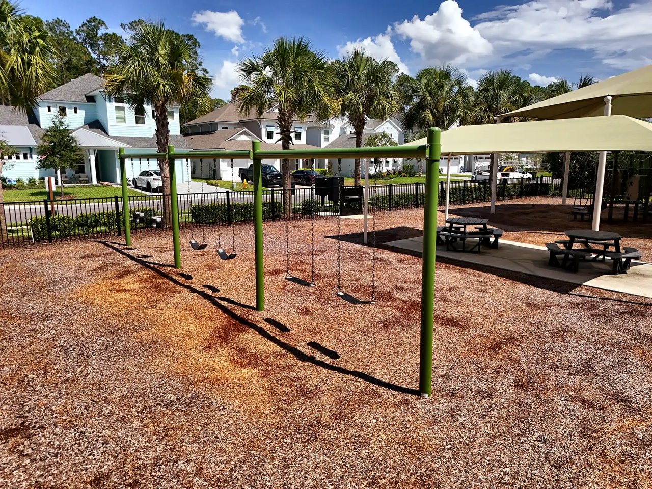 Swing set and shaded picnic tables in a fenced playground, with Palm Crest homes and palms behind.