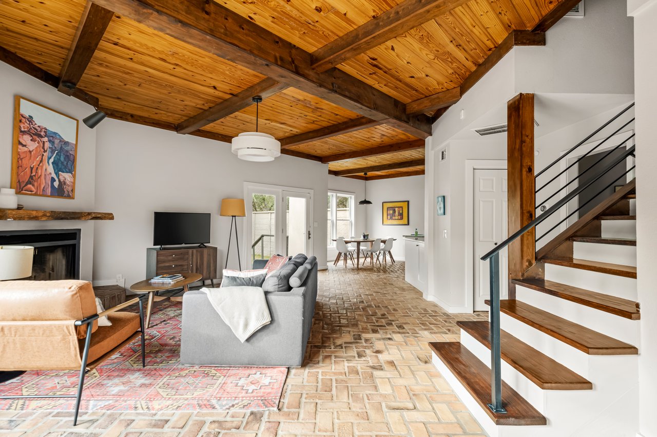 Open-plan living room with vaulted timber ceiling, brick flooring, and staircase beside the dining area.