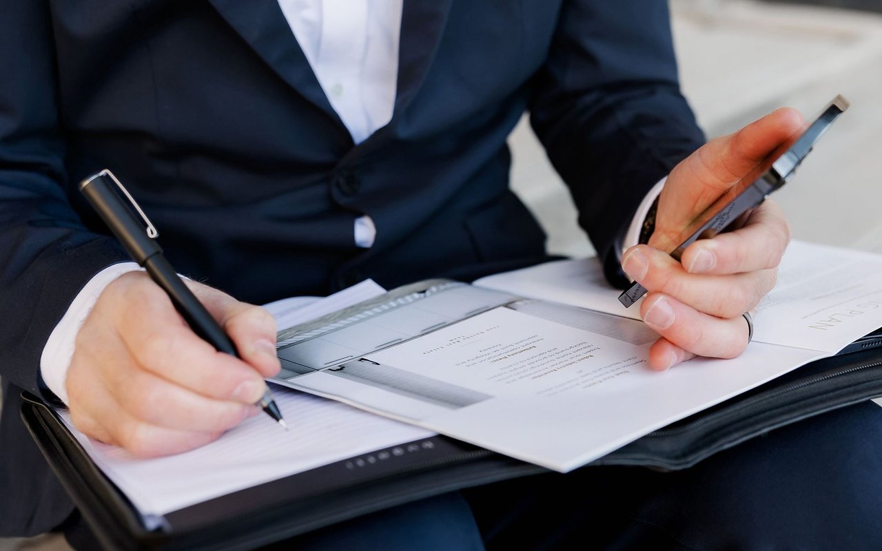 A person in a dark suit signs paperwork while holding a pen over the documents.