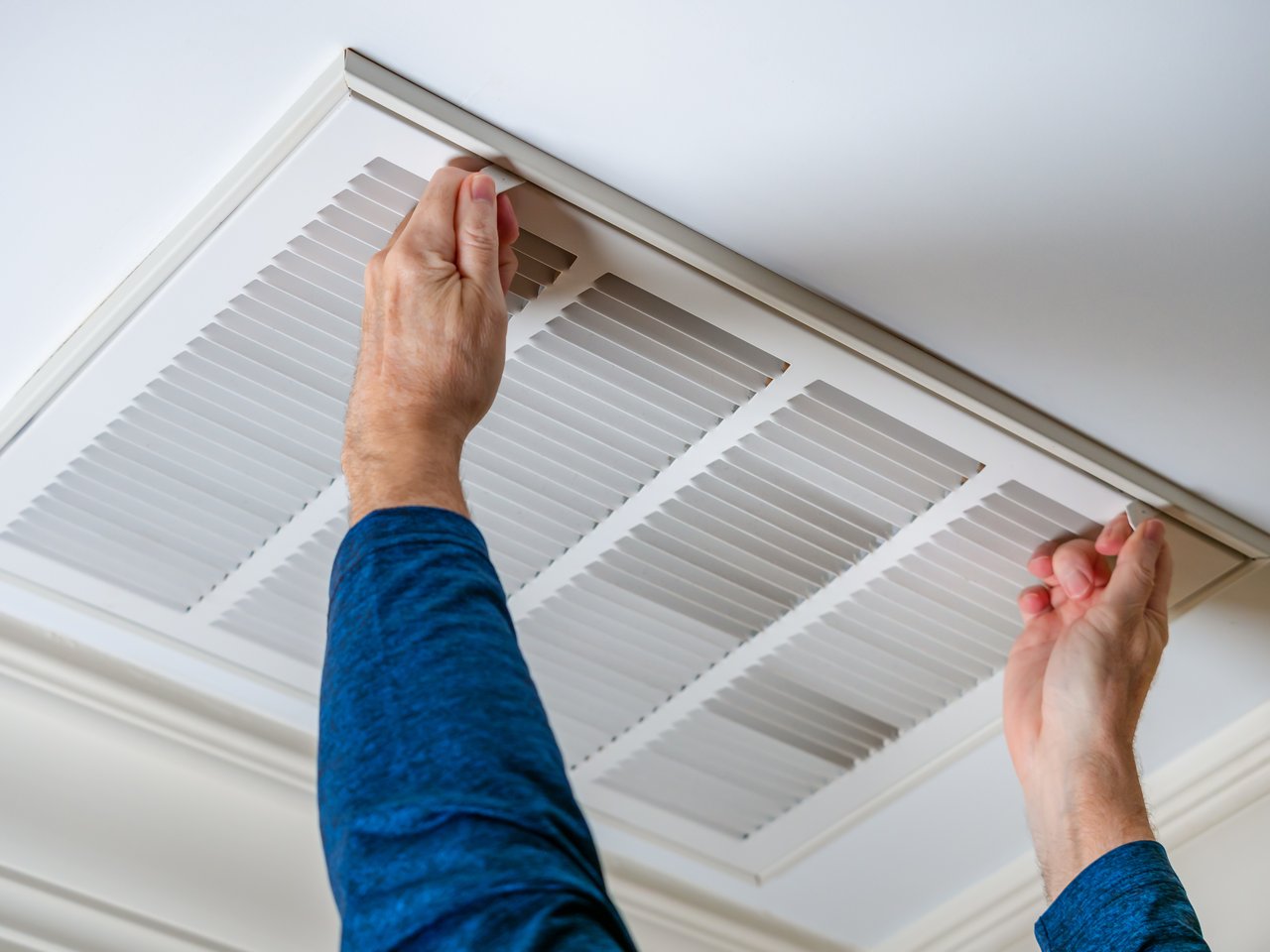 A person wearing a blue shirt, perhaps a real estate agent, adjusts an air vent cover on the ceiling.