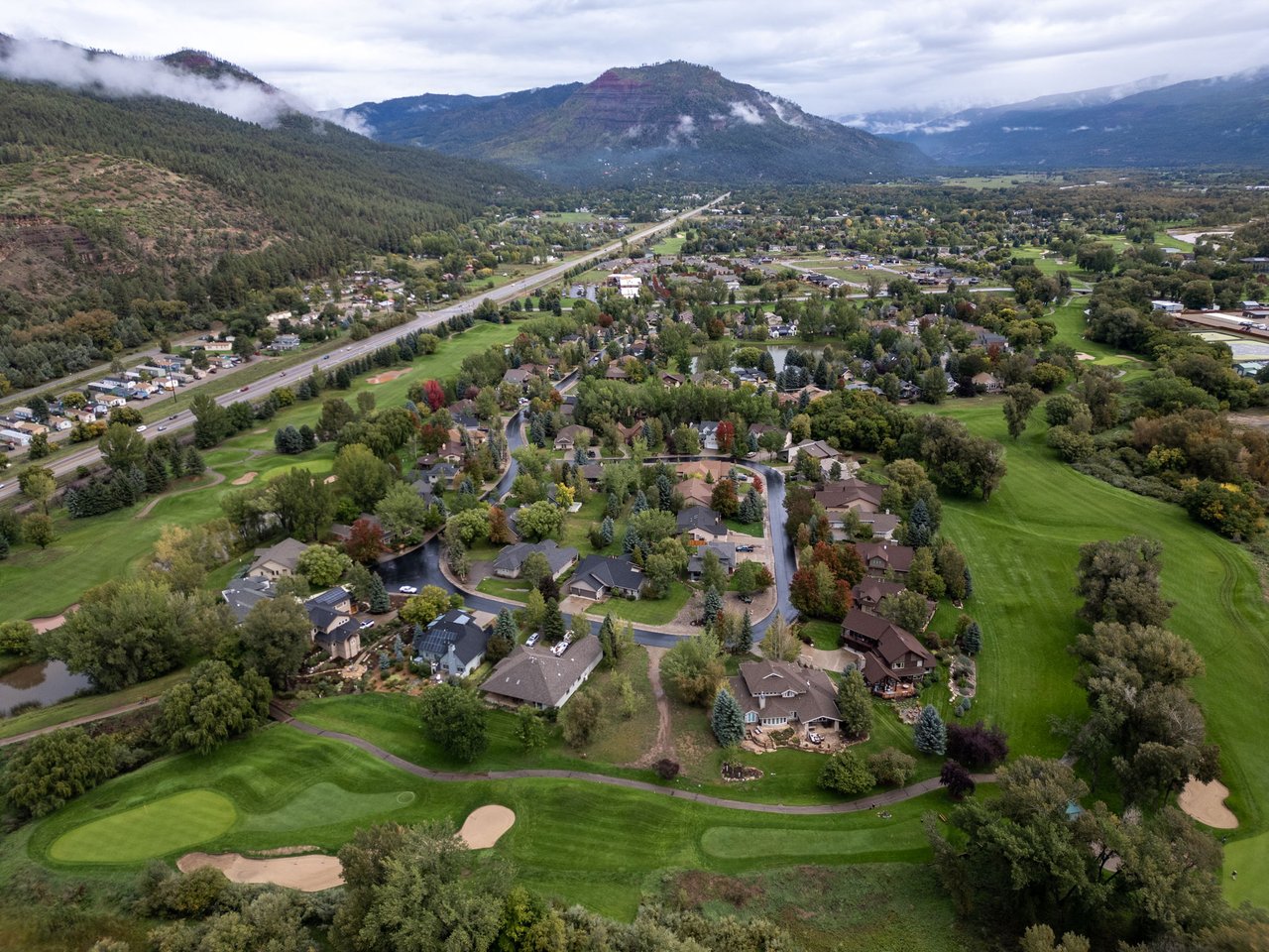 Aerial neighborhood beside a golf course, with homes, a highway, and misty mountains beyond.