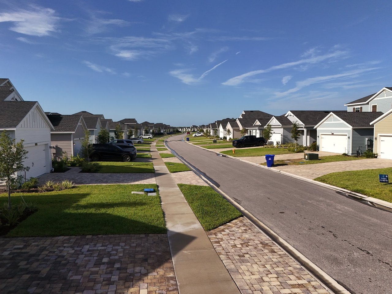 Residential street lined with detached homes, driveways, and lawns beneath a blue sky.