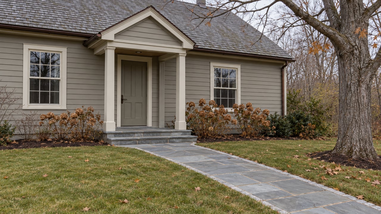 Weathered gray wood-clad home with a sheltered entry, bluestone walk, and deep late-autumn lawn beneath bare maple branches.