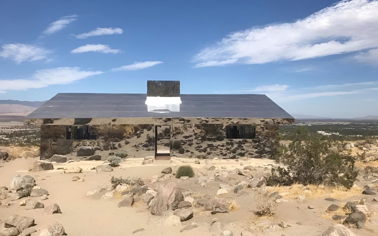 Mirrored house art installation in a rocky desert landscape beneath a blue sky.