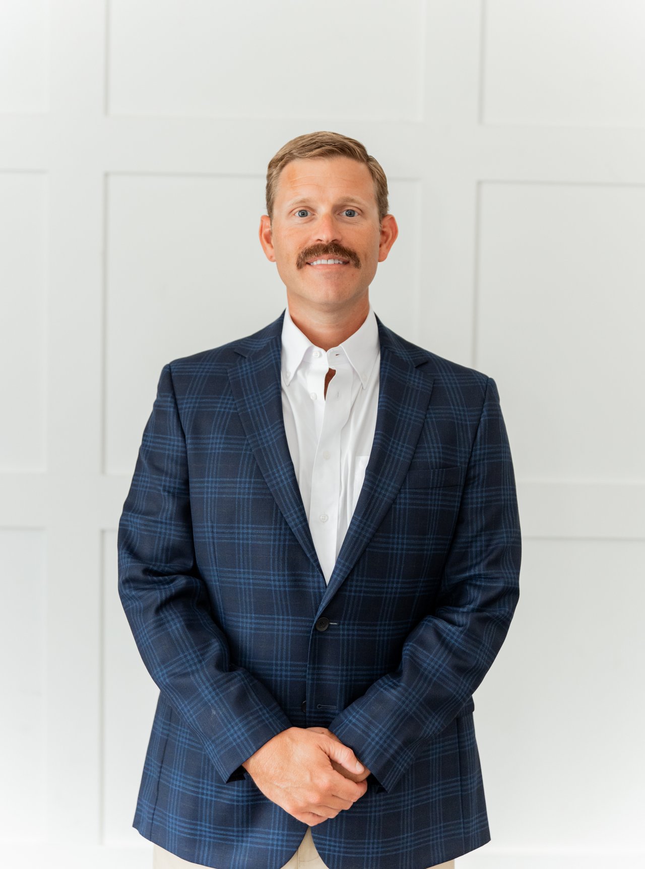 Eddie Kendall in a navy plaid suit, standing with his hands clasped against a white paneled wall.