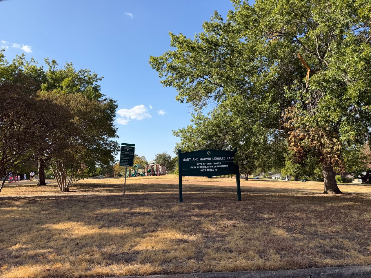 Mary and Marvin Leonard Park sign beneath shade trees, with a playground in the background.
