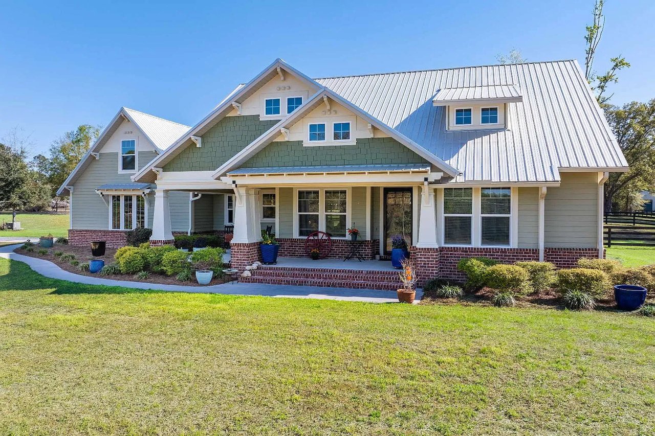 Country-style home with a silver metal roof, covered brick porch, landscaped beds, and broad lawn.