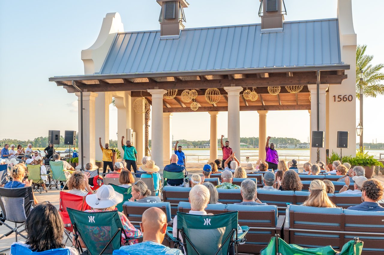 Crowd watching a live performance at a waterfront pavilion with a blue roof.