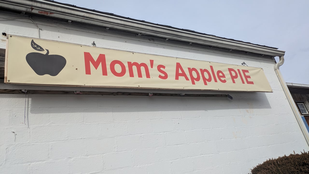 A banner for "Mom's Apple Pie" with a black apple logo on a white brick building at Hill High Marketplace in Round Hill, VA.