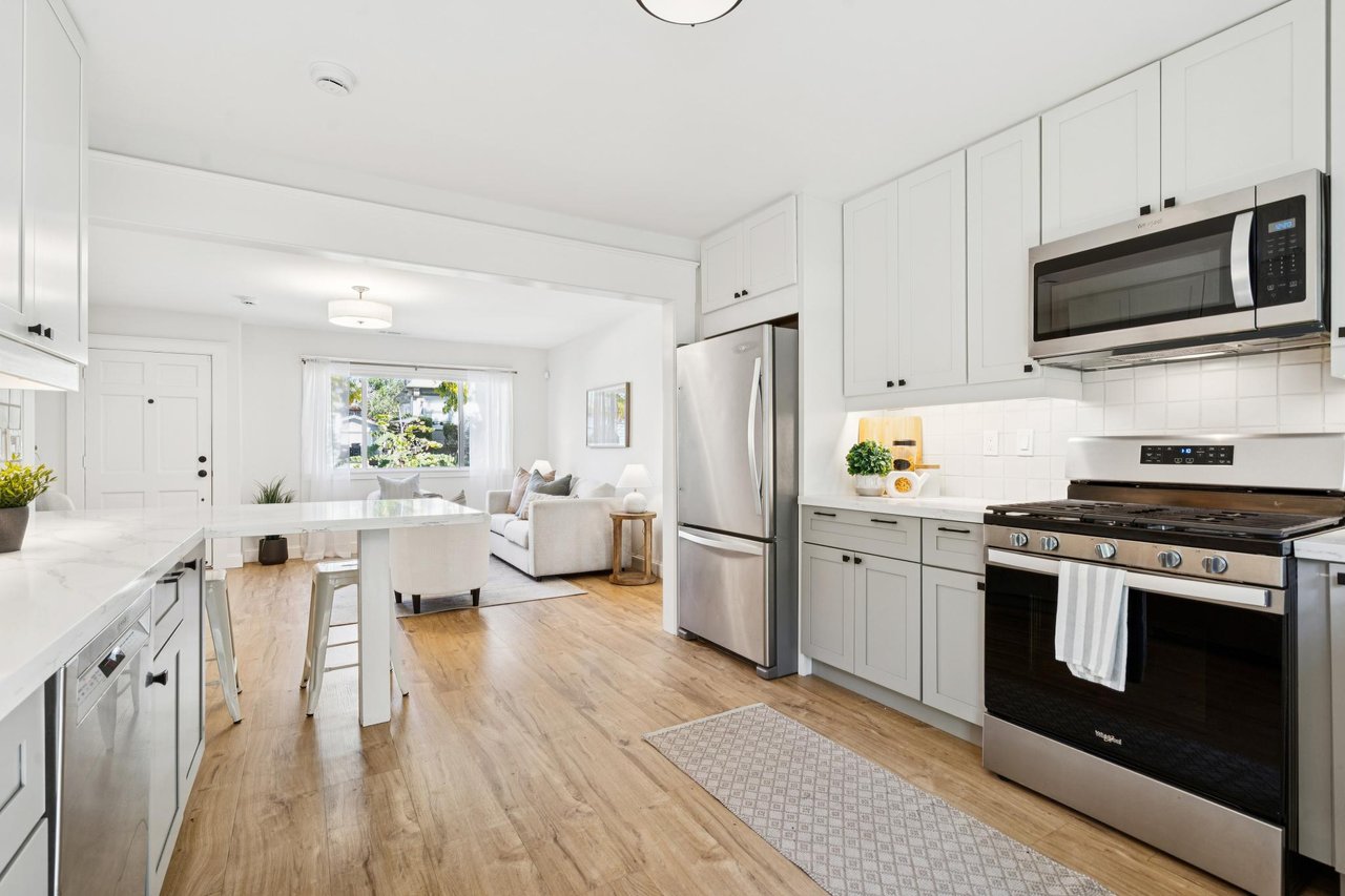 Bright white kitchen with stainless steel appliances and breakfast island opening to a living room.