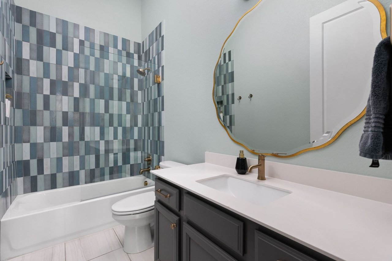 Bathroom with blue-gray tiled tub surround, dark vanity, white countertop, and gold-framed mirror.