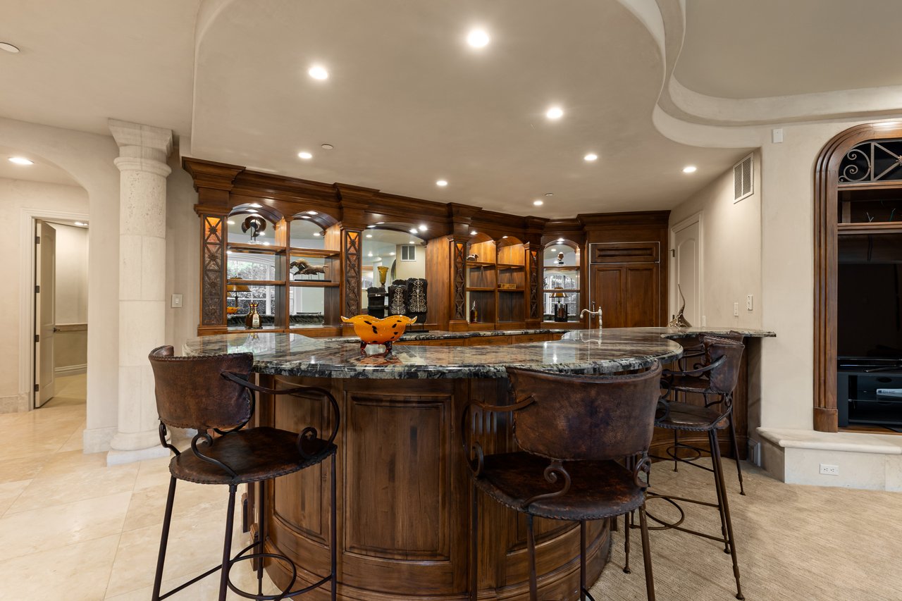 Furnished home bar with a curved stone-topped island, leather stools, and dark wood cabinetry.