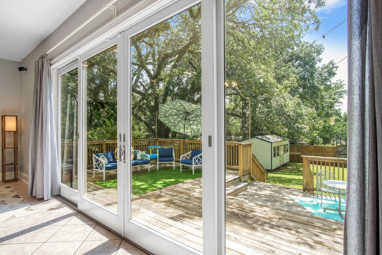 A bright living room with large glass sliding doors opens to a wooden deck. Outside, there's seating, lush greenery, and a clear blue sky. Calm and inviting.