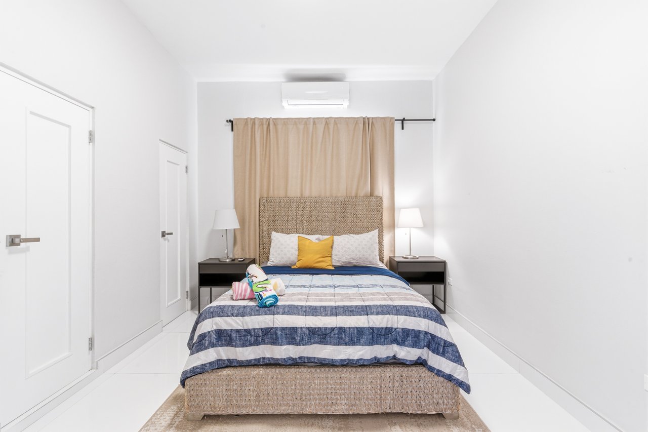 Furnished bedroom with a striped bed, woven headboard, beige curtains, and wall-mounted air conditioner.