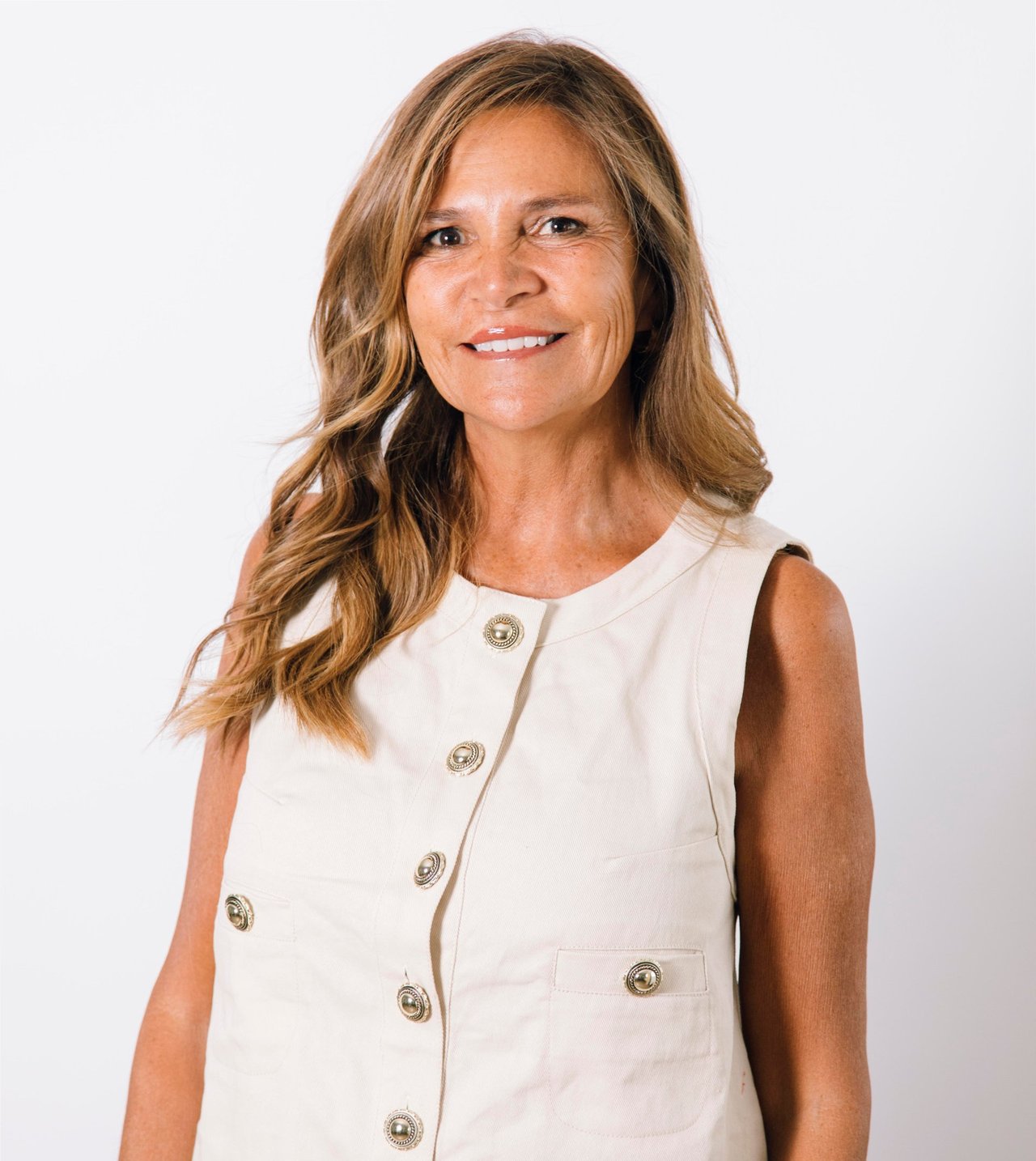 Smiling woman with long highlighted hair wearing an ivory vest with ornate buttons.