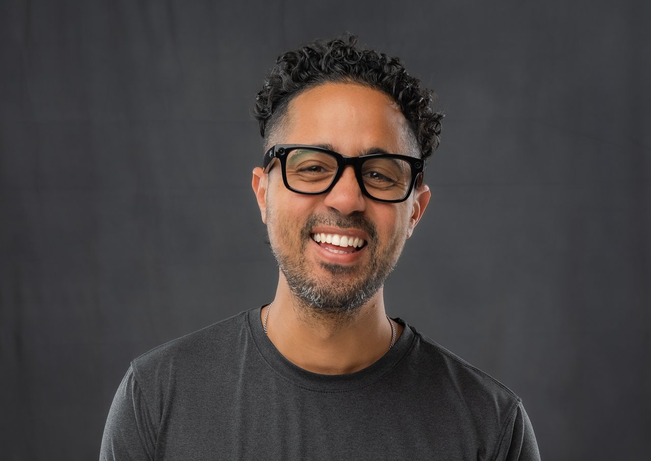 Smiling man with curly hair and black glasses against a dark backdrop.