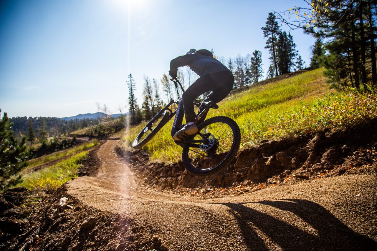 A mountain biker catching air off a dirt berm on a dedicated singletrack trail under a bright blue sky.