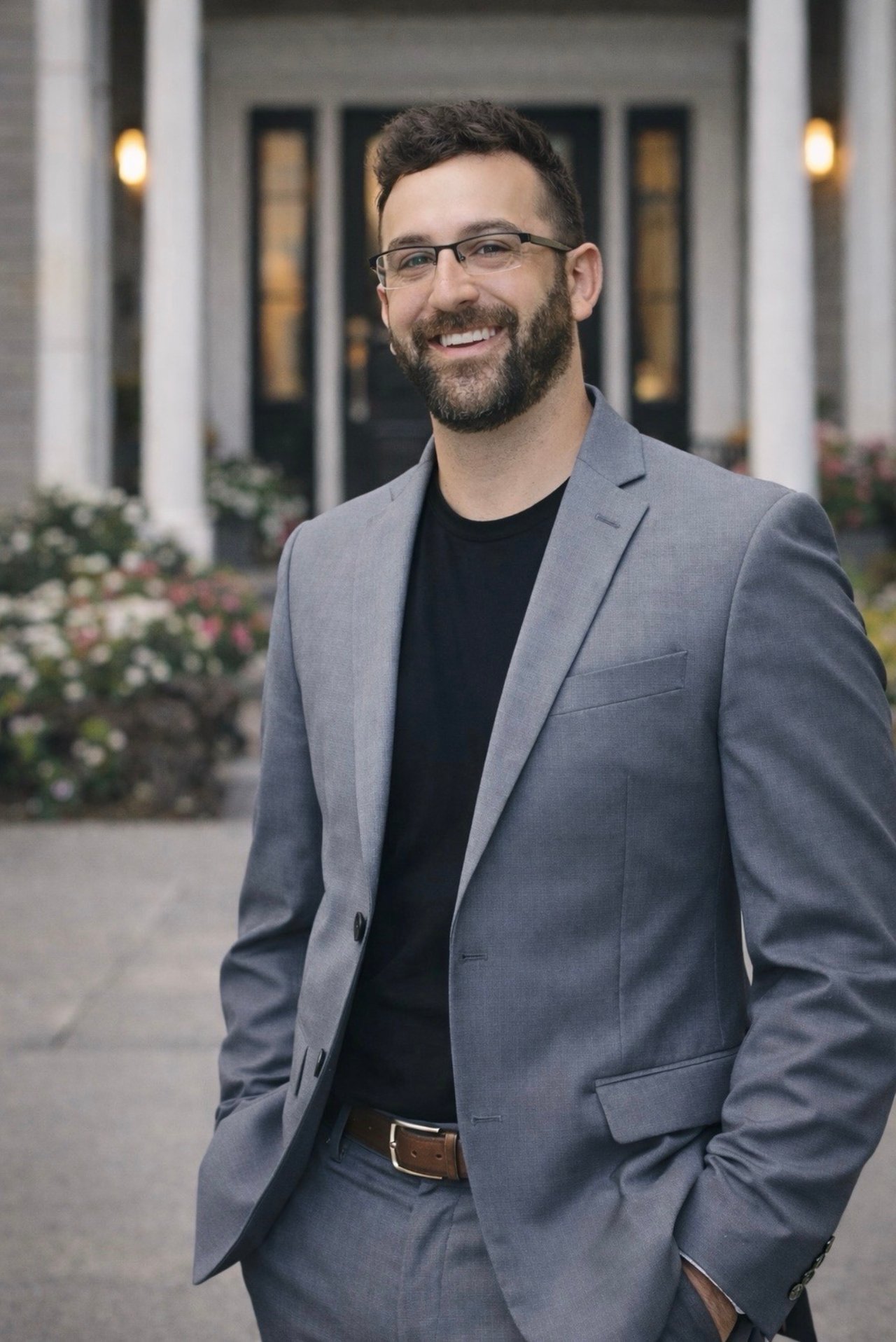 Derek Crippin smiling in glasses and a gray suit outside a building.