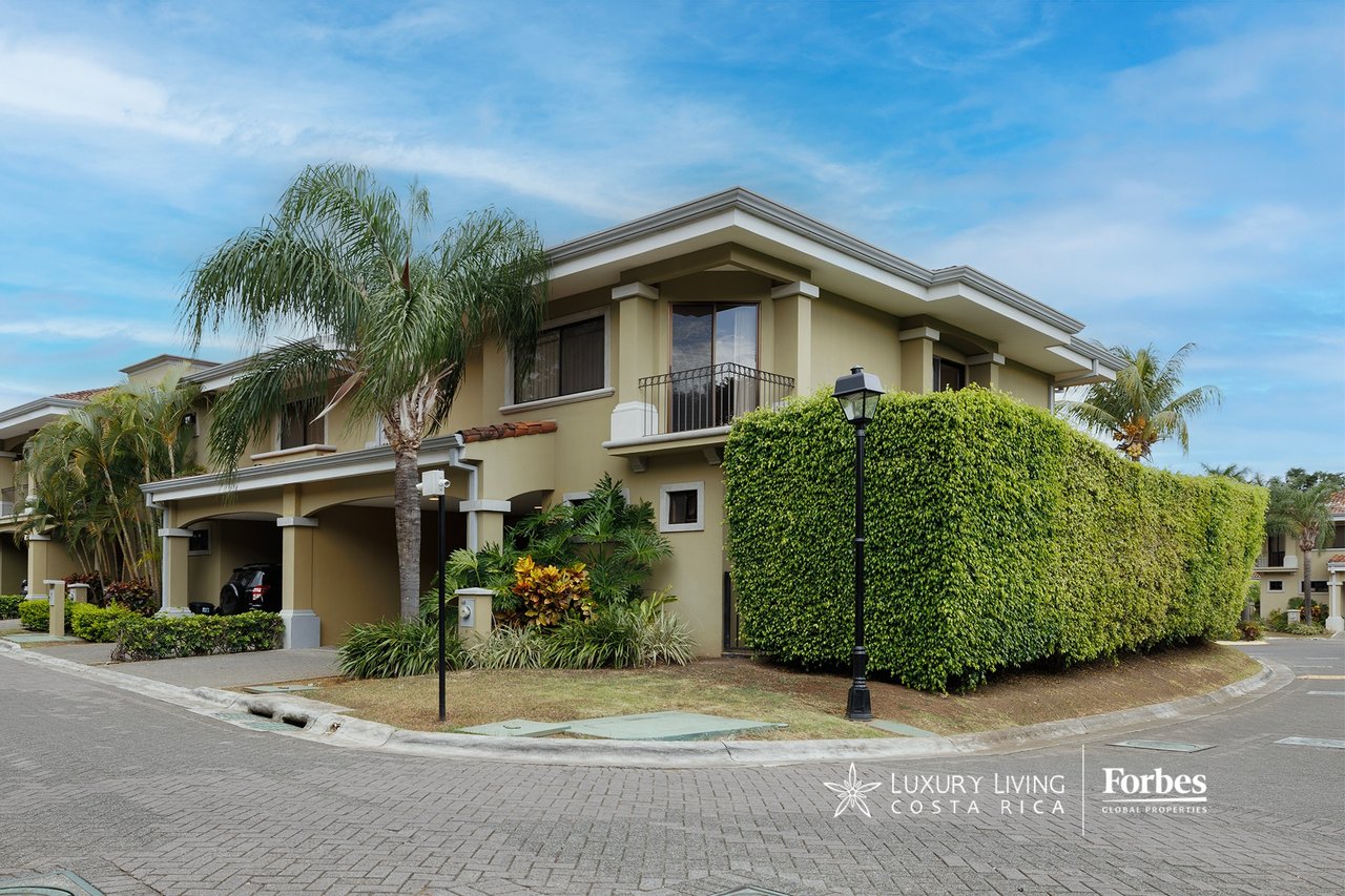 Corner home with a covered carport, balcony, palm trees, and a tall trimmed hedge.