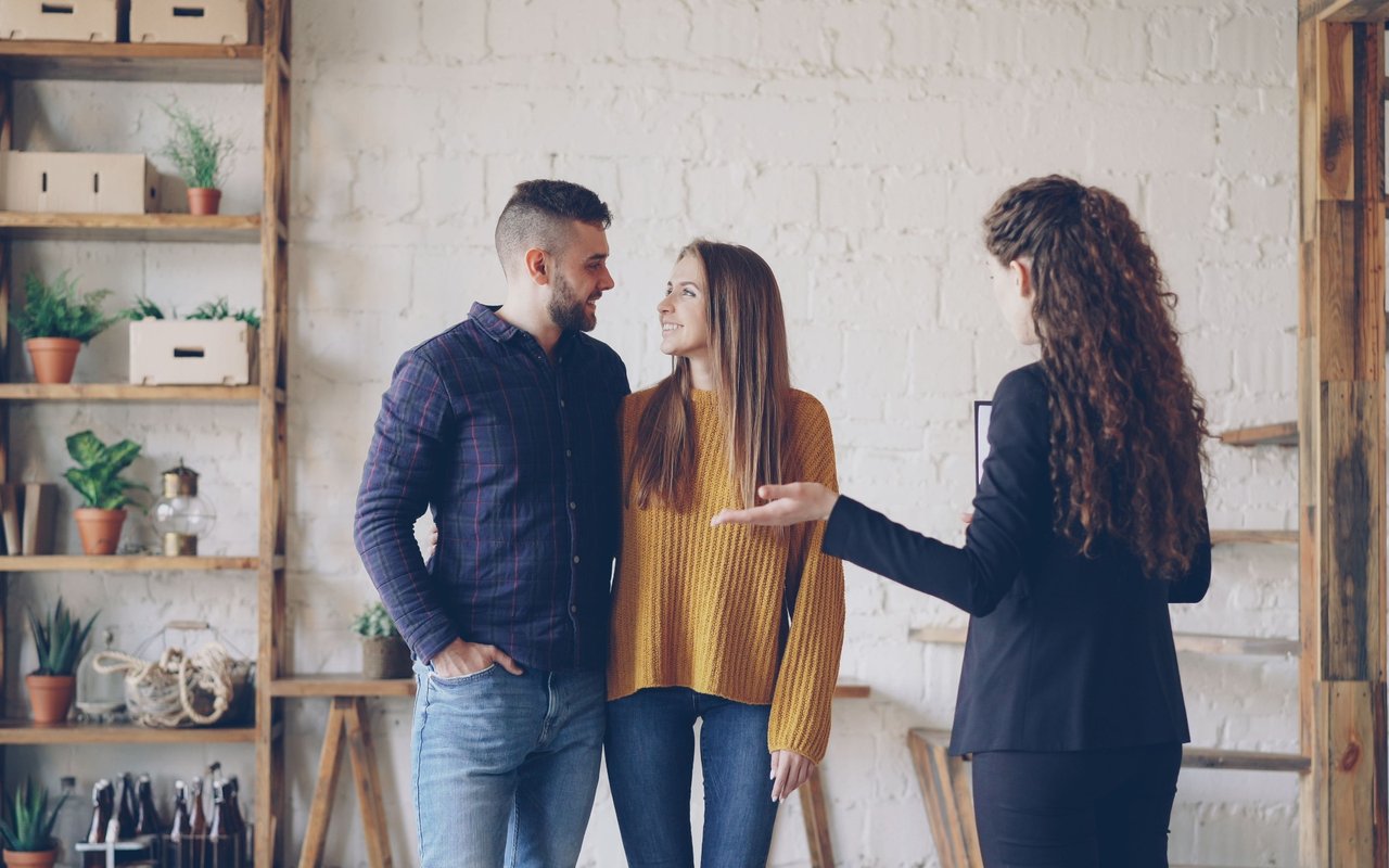 A real estate agent talks with a smiling couple during a home viewing in a bright room.