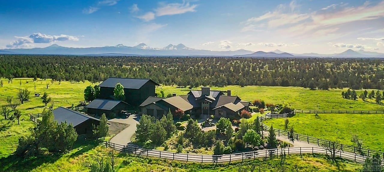 Aerial view of a ranch-style home amid green fields, with the Cascade Mountains beyond.