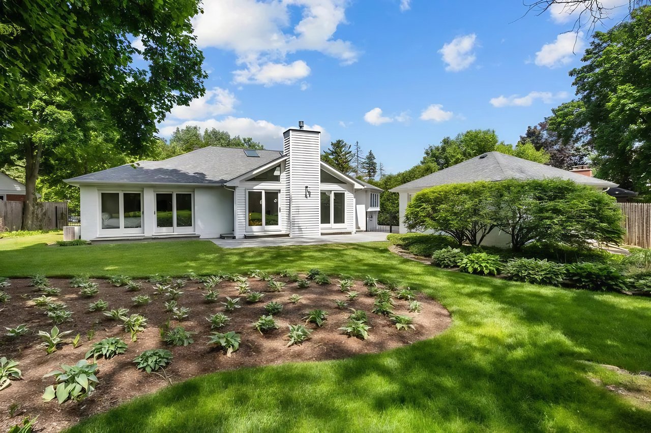 White single-story home with a prominent chimney, landscaped garden beds, and mature trees in the backyard.