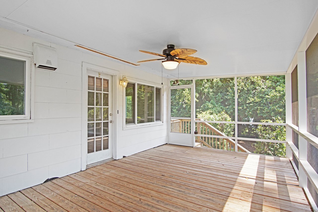 Covered porch with wooden flooring, white walls, and a ceiling fan. Screened windows reveal lush green trees outside, creating a serene, airy ambiance.