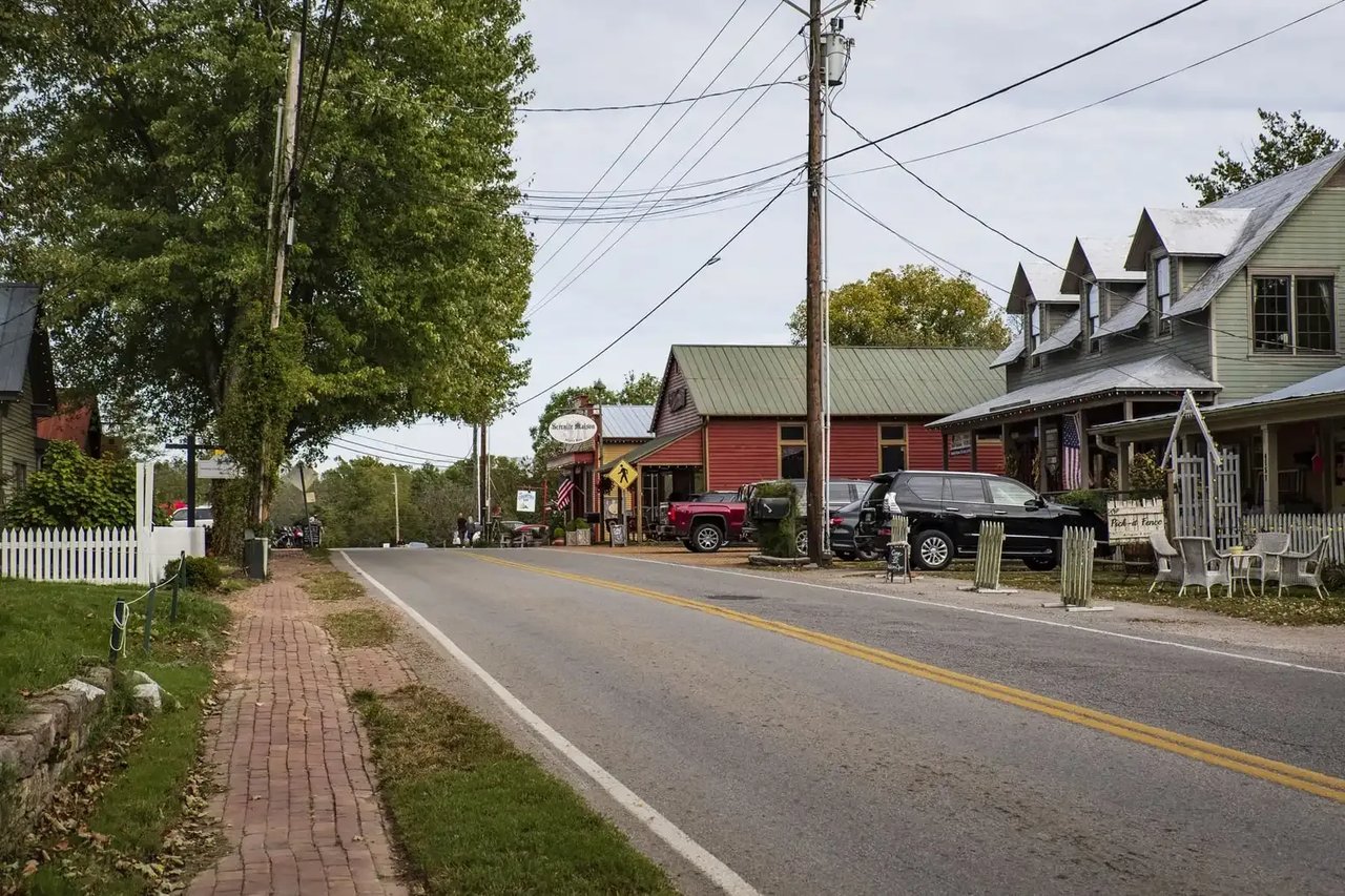 Small-town street with a red storefront, roadside shops, parked vehicles, and a brick sidewalk.
