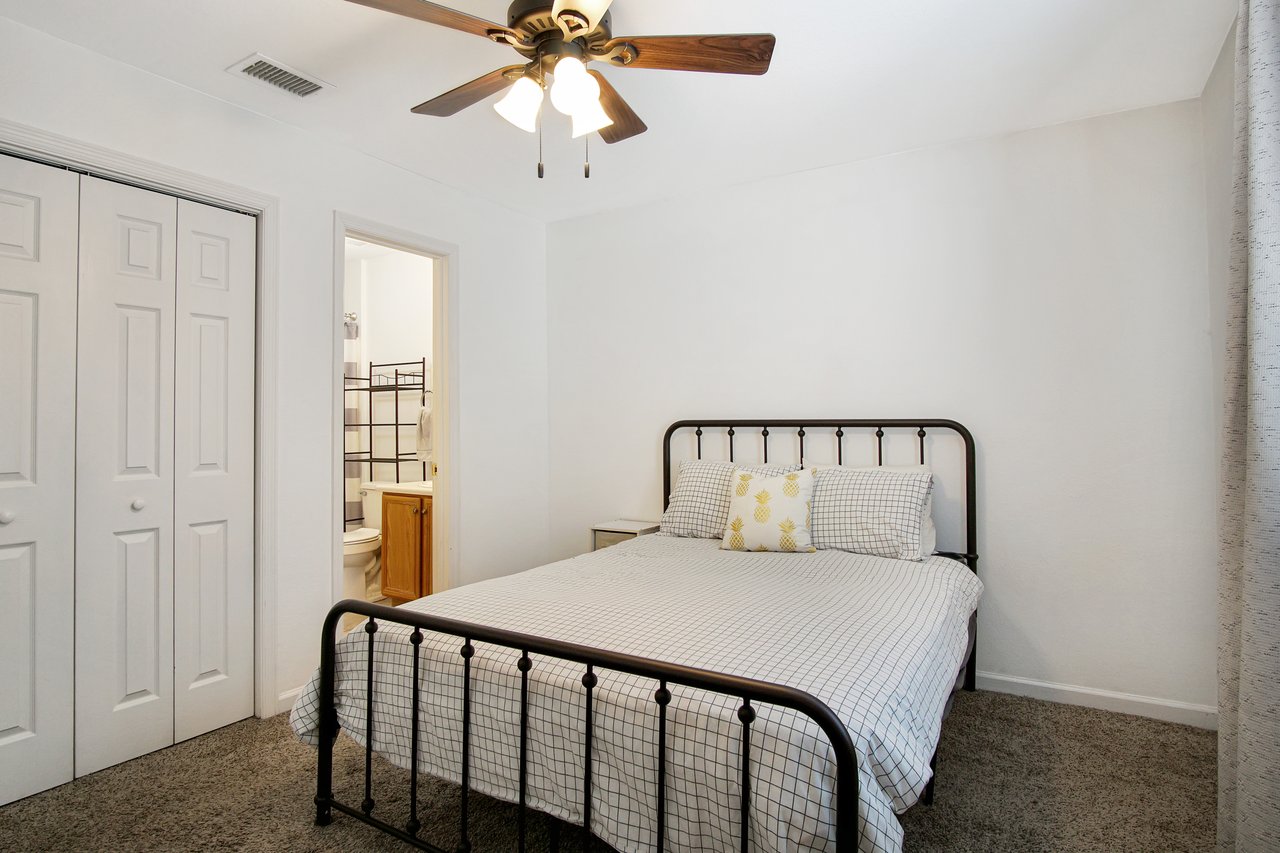 Cozy bedroom with a double bed, plaid bedding, and pineapple-patterned pillows. A ceiling fan and adjacent bathroom add functionality and comfort.