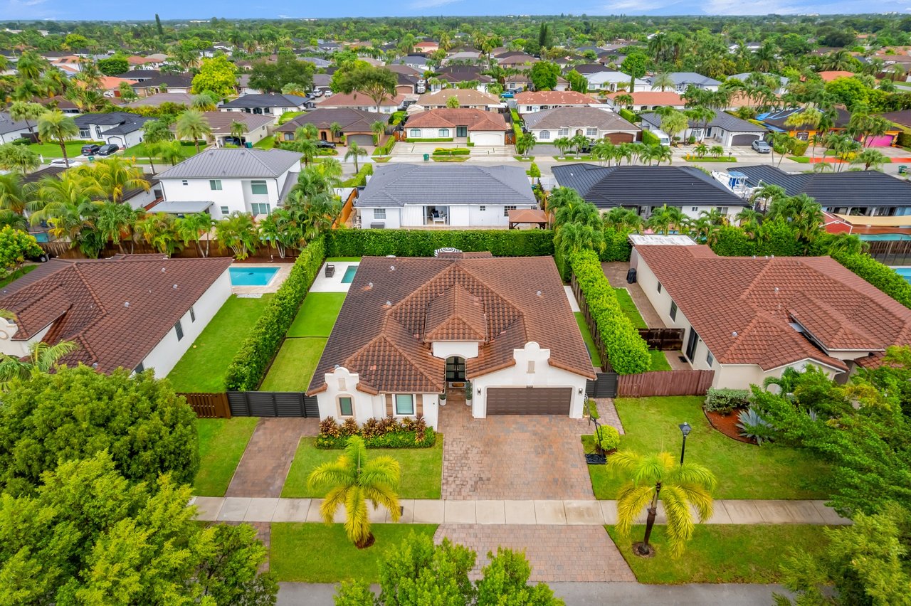 Aerial view of a tile-roofed home with a paver driveway and backyard pool among palm-lined houses.