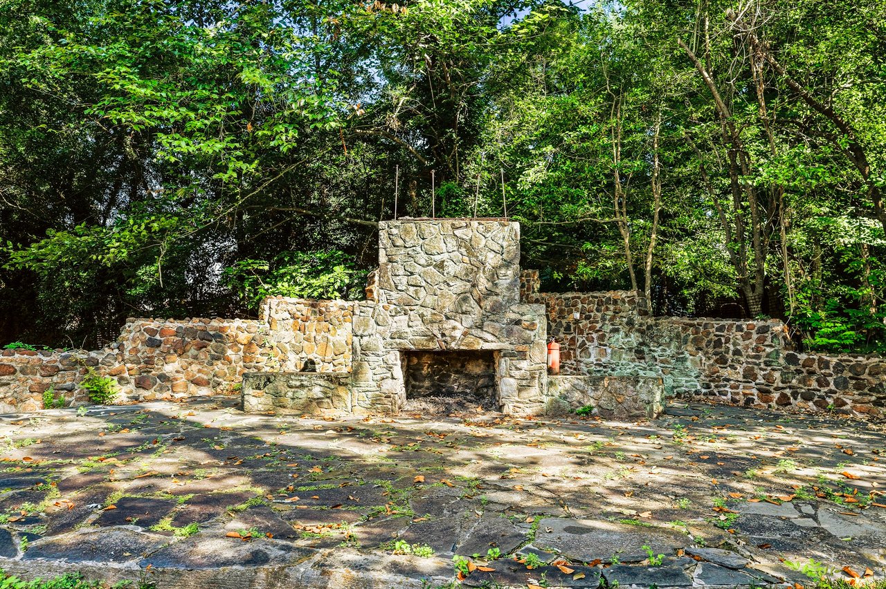 Stone outdoor fireplace and walled patio beneath leafy trees.