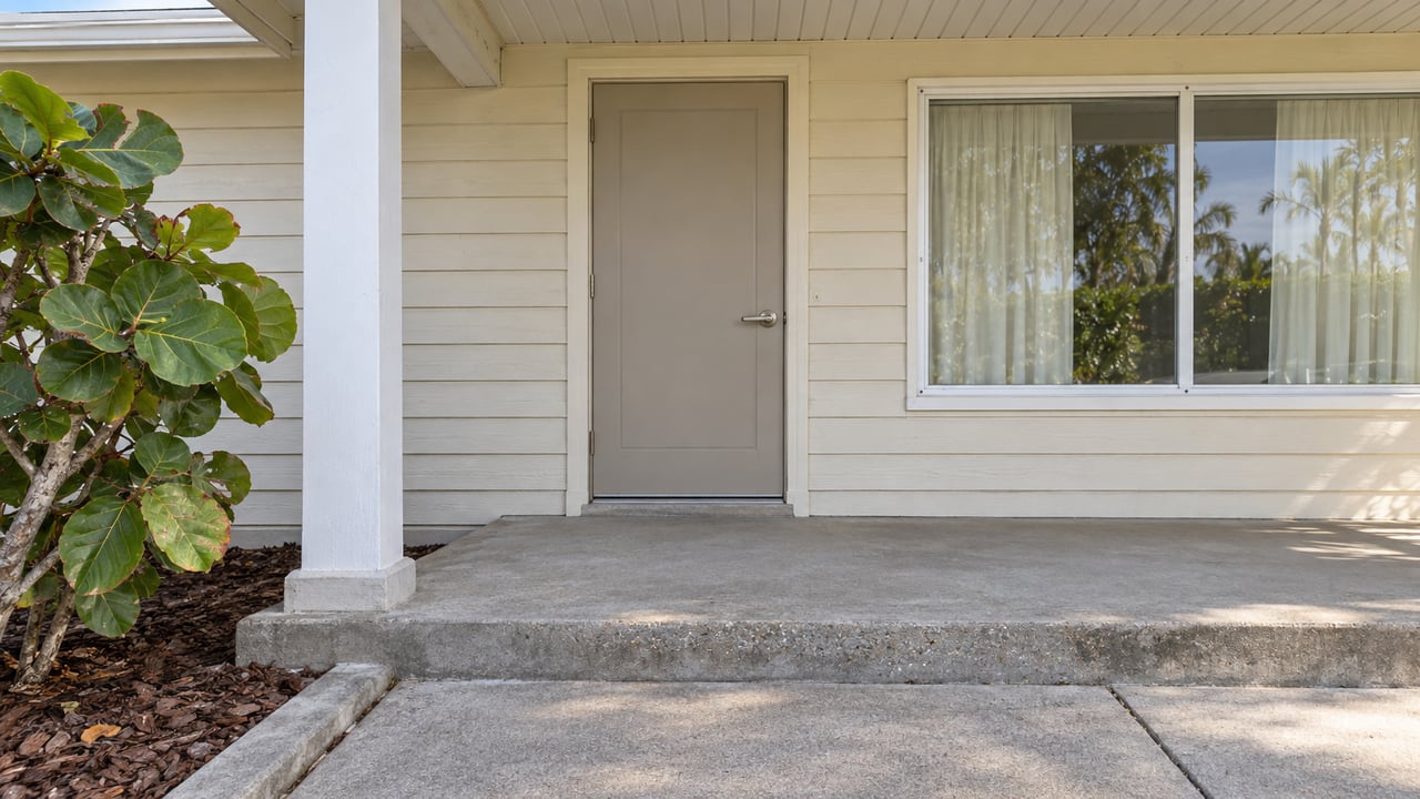 Cream-sided ranch entrance with a white porch post, closed beige door, picture window and concrete step beside sea-grape leaves.