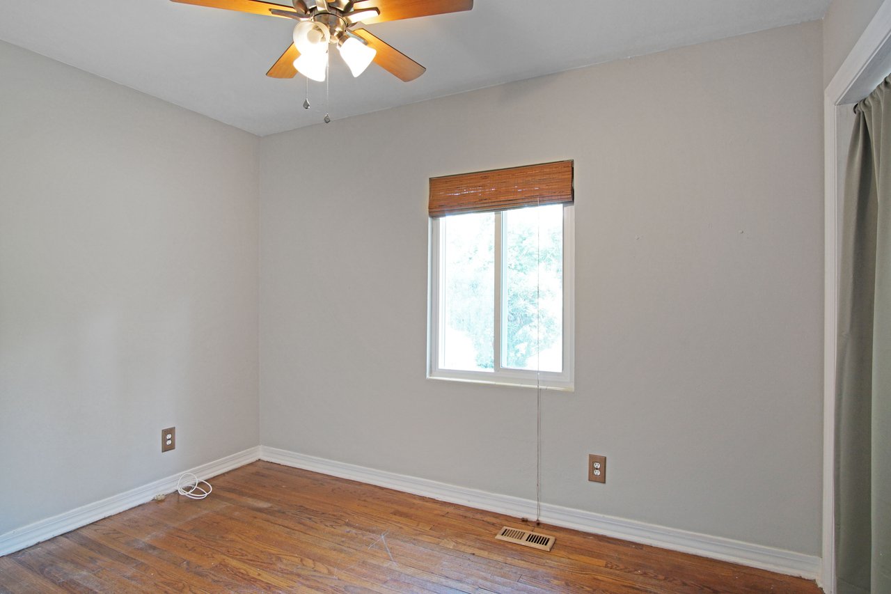 Empty room with light gray walls, wooden floor, a window with bamboo blinds, and a ceiling fan with lights. The space feels simple and clean.