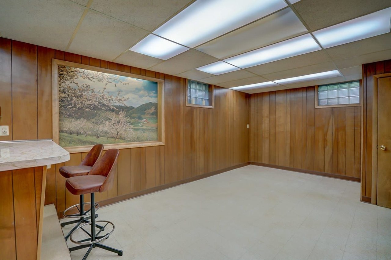 Wood-paneled basement room with a built-in counter, two bar stools, and fluorescent ceiling lights.