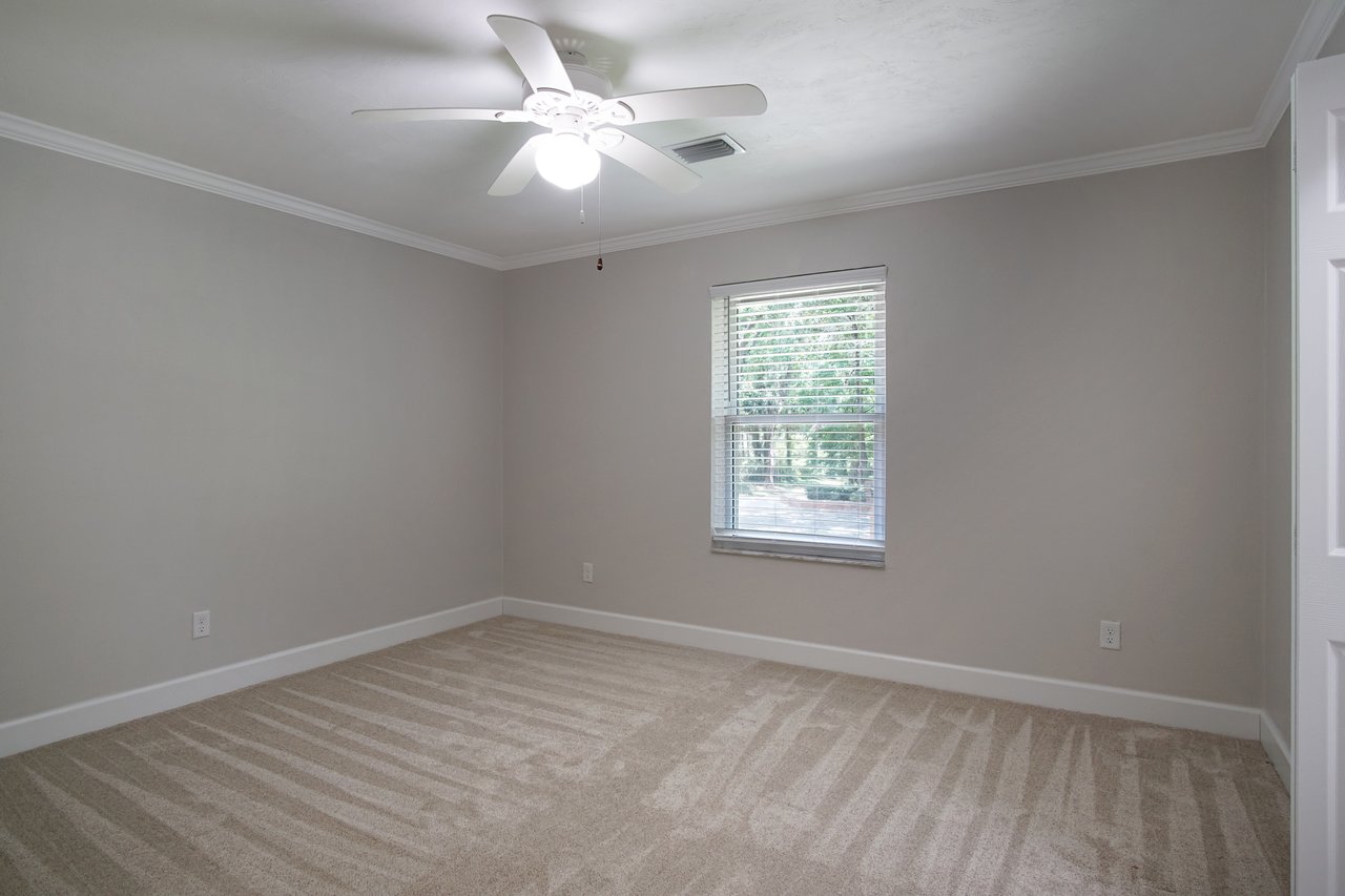 A beige, empty room with a light carpet, a ceiling fan with a bright light, and a window with blinds. Soft natural light creates a calm, inviting atmosphere.