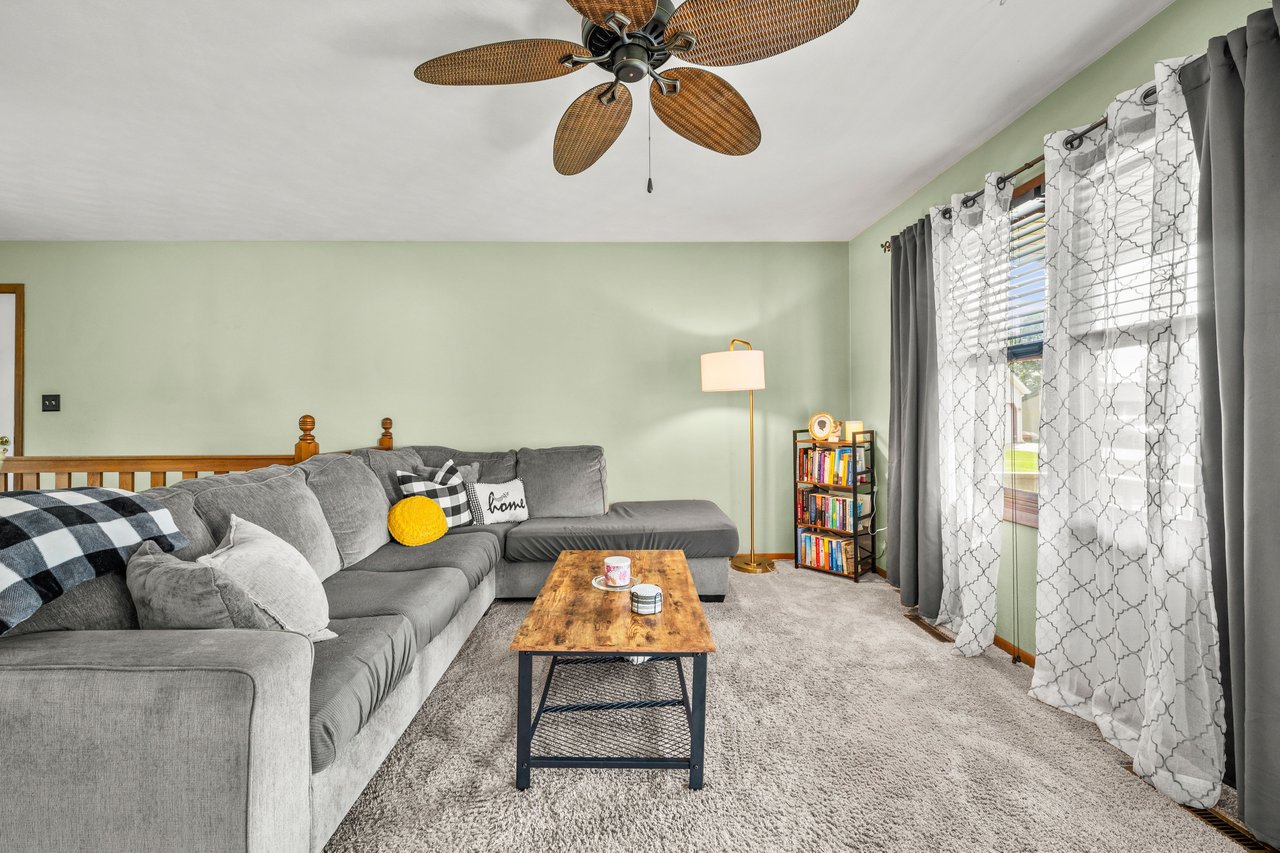 Living room with a gray sectional, wood coffee table, patterned curtains, and woven-blade ceiling fan.