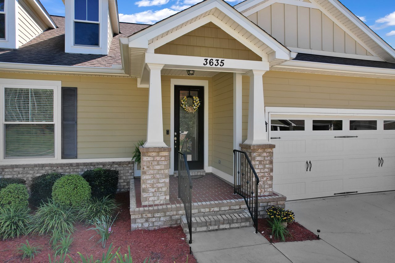 A welcoming yellow house with white trim, featuring a brick porch with two columns and a black front door adorned with a wreath. Adjacent is a white garage door. Bushes and flowers in the garden add charm under a bright blue sky.